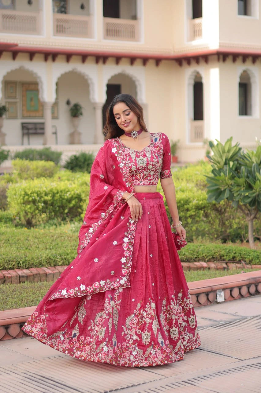 Woman in a pink embroidered traditional outfit standing in front of a building with greenery.